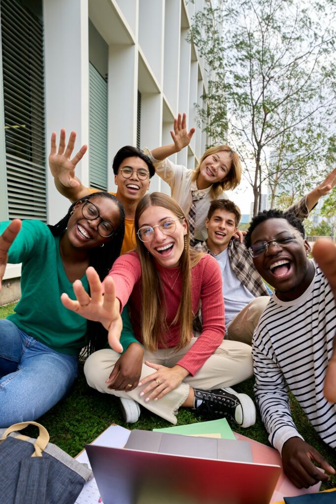 Amusing portrait of group diverse young friends excited waving at camera with hands sitting on grass